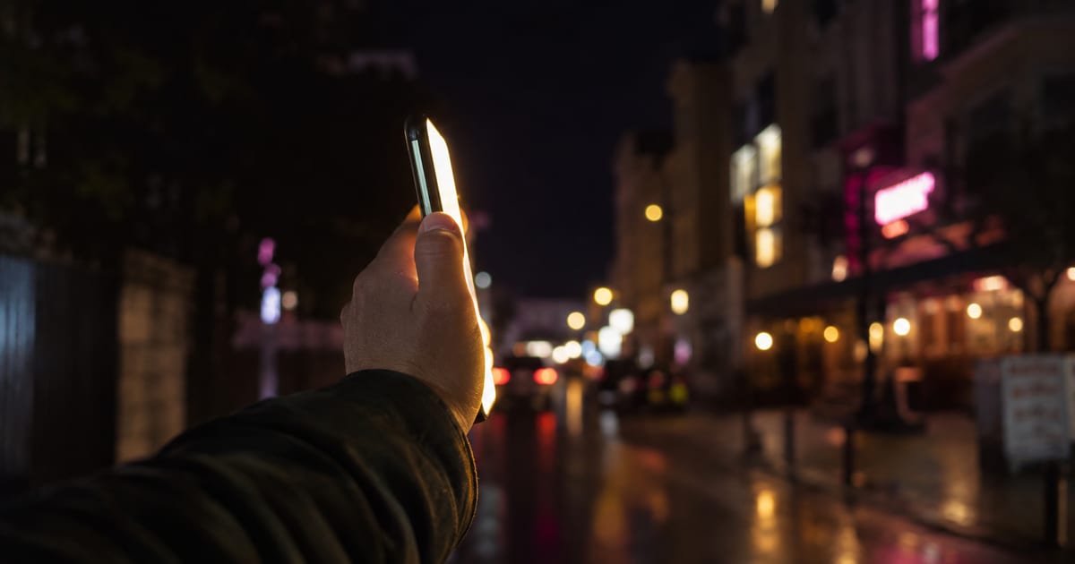 A phone held up at arm's length in colored evening light on a city street, its screen glowing against a dark background
